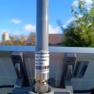 Op de afbeelding: Een grijze glasvezelantenne met een zilveren connector is bevestigd aan een grijze basis. De antenne heeft een label met de tekst "Fiberglass Antenna", modelinformatie, frequentie en versterking. De achtergrond toont een blauwe lucht en daken.