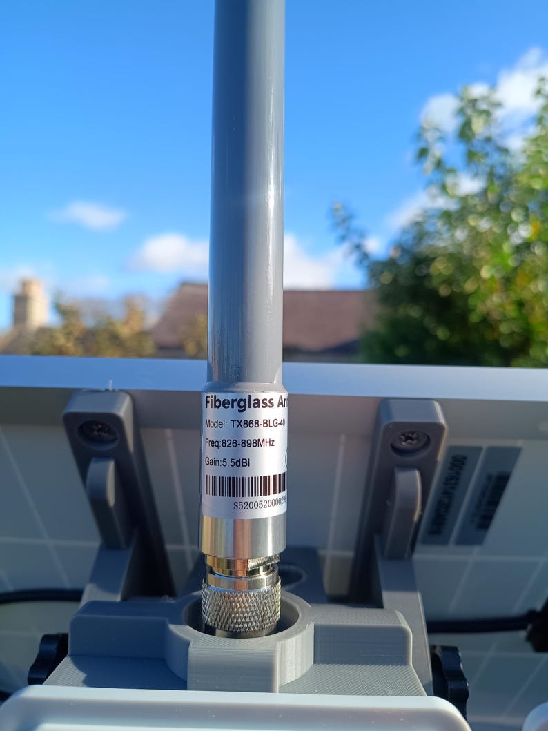 Op de afbeelding: Een grijze glasvezelantenne met een zilveren connector is bevestigd aan een grijze basis. De antenne heeft een label met de tekst "Fiberglass Antenna", modelinformatie, frequentie en versterking. De achtergrond toont een blauwe lucht en daken.