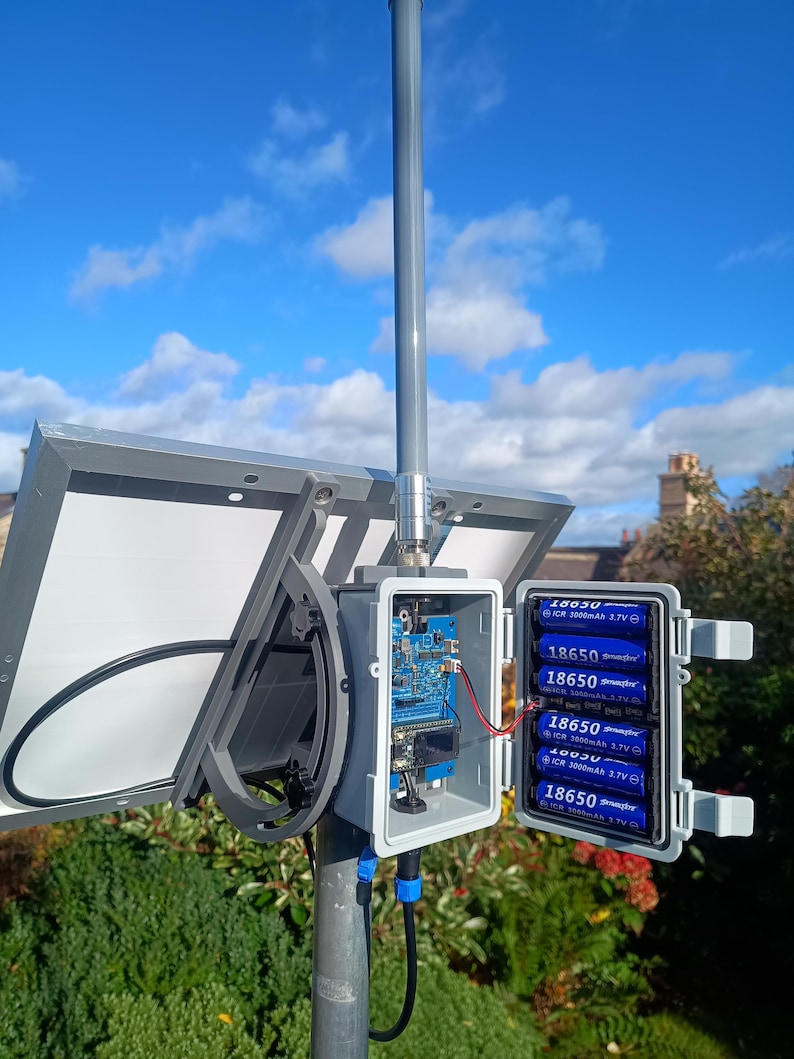 Op de afbeelding: Een zonnepaneelsysteem gemonteerd op een paal, met een grijze behuizing met een printplaat en zes 18650 3,7V batterijen. Het systeem omvat een zonnepaneel en een blauwe en zwarte kabel. De achtergrond toont een blauwe lucht met wolken en groene bladeren.