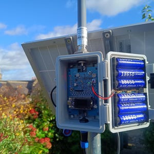 Op de afbeelding: Een zonnepaneelsysteem met een open grijze doos met een printplaat en zes blauwe 18650-batterijen. Het systeem is gemonteerd op een metalen paal, met een blauwe lucht en vegetatie op de achtergrond.