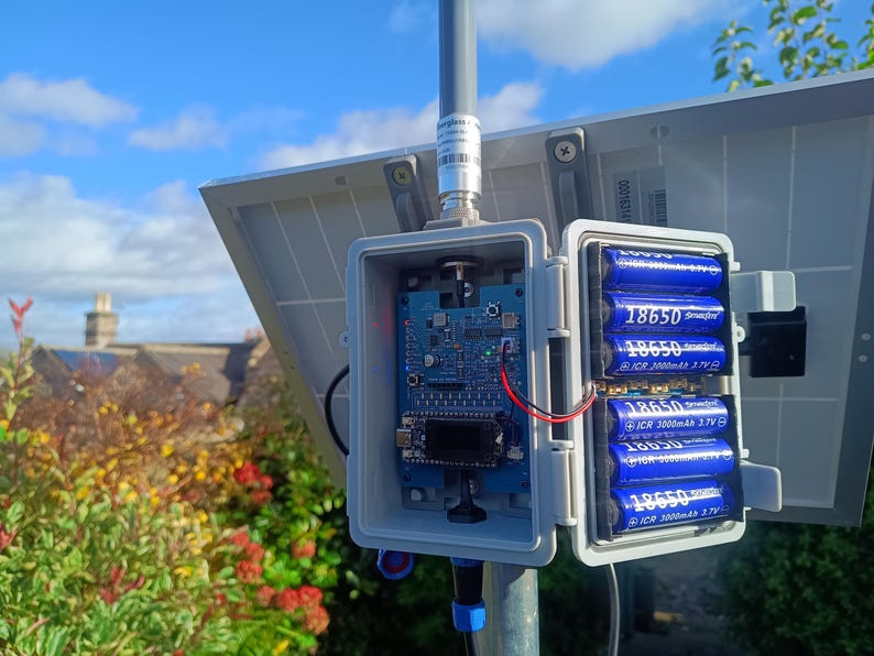 Op de afbeelding: Een zonnepaneelsysteem met een open grijze doos met een printplaat en zes blauwe 18650-batterijen. Het systeem is gemonteerd op een metalen paal, met een blauwe lucht en vegetatie op de achtergrond.