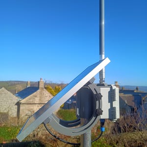 Op de afbeelding: Een zonnepaneel gemonteerd op een metalen paal, schuin omhoog. Het paneel is zilverkleurig met een donkergrijze beugel en een grijze doos. Een blauwe kabel is aangesloten op de doos. De achtergrond toont een heldere blauwe lucht en daken.