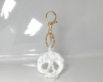 Puede incluir: Un llavero de calavera de ganchillo blanco con un cierre y un anillo dorados. La calavera tiene un diseño detallado con cuencas de los ojos y nariz visibles. Pequeños diamantes de imitación amarillos se colocan en la parte inferior de la calavera.