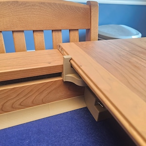 May include: A close-up of a wooden table with a leaf extension. The leaf is raised and supported by a metal bracket. The table is made of light wood with a natural finish.