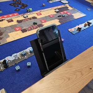 May include: A blue felt game table with a board game set up on it. There are dice, cars, and cards on the table. A smartphone is propped up in a stand on the table. The table is made of wood.