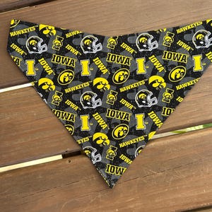 May include: A triangular pet bandana featuring a repeating pattern of the Iowa Hawkeyes logo, including the team name, helmet, and the word "IOWA" in yellow on a gray background. The bandana is made of fabric.