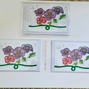 May include: Three blank greeting cards with a watercolor painting of purple and pink flowers on each. The flowers are surrounded by green leaves and a green ribbon.