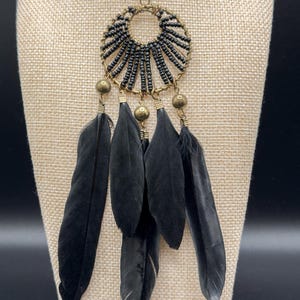 May include: Black feather earrings with a circular beaded design. The earrings feature a gold-colored metal frame and accents, with multiple black feathers hanging below. The earrings are displayed on a beige surface.