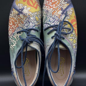 May include: Pair of canvas shoes with a colorful, vintage-style design featuring text and illustrations. The shoes have blue laces and a cream-colored interior. The design includes the words "LA PAIX" and "DERNE".