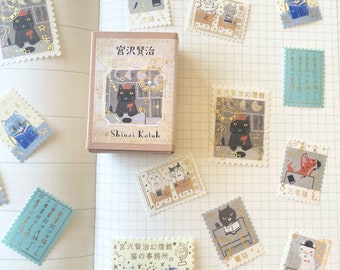 May include: A set of 12 decorative stickers featuring illustrations of cats in various poses and settings. The stickers are printed on a white background with gold accents and feature Japanese text. The stickers are packaged in a small box with a brown background and gold lettering. The text on the box reads "宮沢賢治" and "Shinzi Katoh".