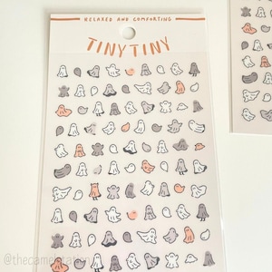 May include: A sheet of stickers featuring a variety of cute ghost illustrations in white, grey, and peach. The stickers are arranged in rows on a white background. The text "TINY TINY" is printed at the top of the sheet. The text "RELAXED AND COMFORTING" is printed above the text "TINY TINY". The text "@thecamelotation" is printed at the bottom of the sheet.