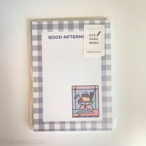 May include: A white notepad with a gray and white checkered border. The notepad has the text "GOOD AFTERNOON" at the top and a small illustration of a dessert in a glass dish with a cherry on top. The notepad also has a small sticker that says "OTE GARU MEMO" and "PAPIER PLATZ".