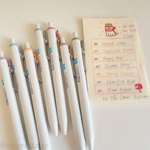 May include: A set of ten white gel pens with colored caps and a notecard with the text "Uni-ball One FIKA 0.5mm" and a list of colors with their names: Avocado Green, Cappuccino Brown, Poppy Red, Sesame Grey, Mango Orange, Nemofila Blue, Plum Purple, by The Camel Station.