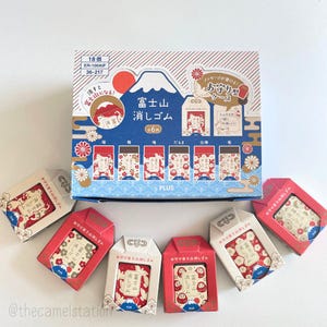 May include: A collection of Japanese eraser sets with Mount Fuji designs. The box is blue and white with red accents, and the individual erasers are in small, decorative boxes. The text on the box reads "18 pieces" and "Eraser".