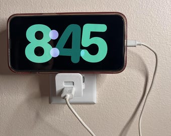 May include: A smartphone displaying the time 8:45 in green numerals, charging via a white cable and adapter plugged into a wall outlet. The phone is in a clear case, mounted on a beige wall.