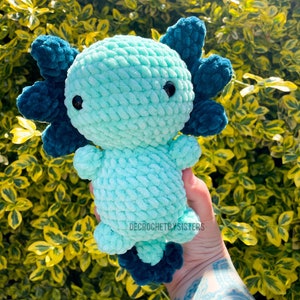 May include: A crocheted axolotl plush toy with a teal body and dark blue fins. The toy is being held in a hand with a tattoo.