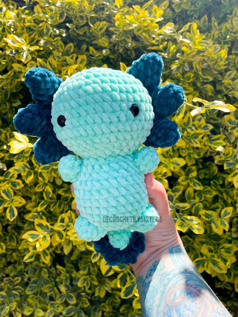 May include: A crocheted axolotl plush toy with a teal body and dark blue fins. The toy is being held in a hand with a tattoo.