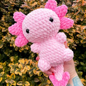 May include: Pink crocheted axolotl plush toy with black eyes. The toy is being held in a hand with a tattoo.