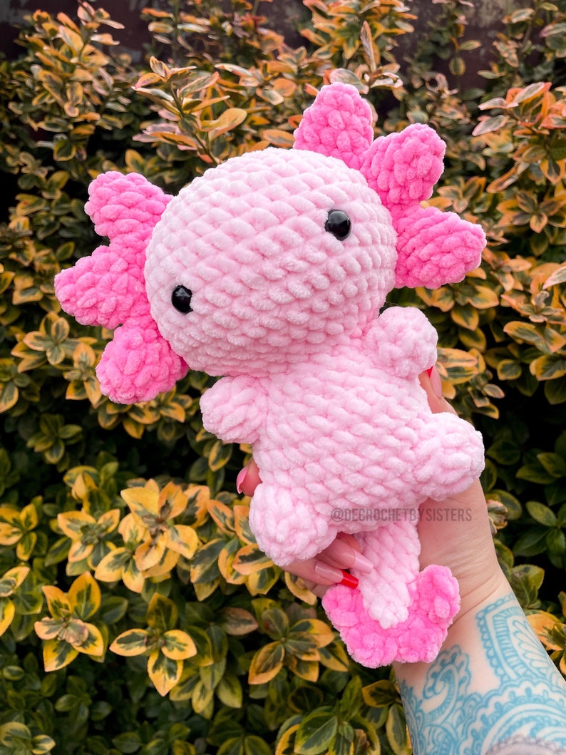 May include: Pink crocheted axolotl plush toy with black eyes. The toy is being held in a hand with a tattoo.