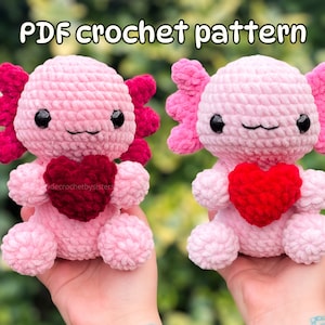May include: Two pink crocheted axolotl plushies holding red heart-shaped crocheted plushies. The axolotls have black eyes and pink and red crocheted details. The plushies are being held by hands.