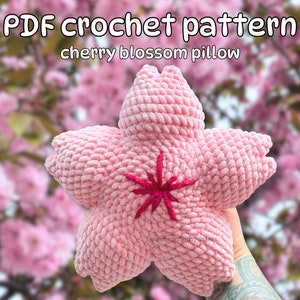 May include: A pink crocheted cherry blossom cushion with a red embroidered centre. The cushion is held in front of a background of pink cherry blossoms.