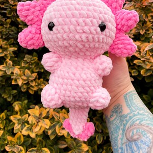 May include: A pink crocheted axolotl plush toy with black eyes. The toy is held in a hand with a tattoo.