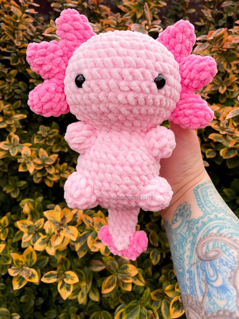 May include: A pink crocheted axolotl plush toy with black eyes. The toy is held in a hand with a tattoo.