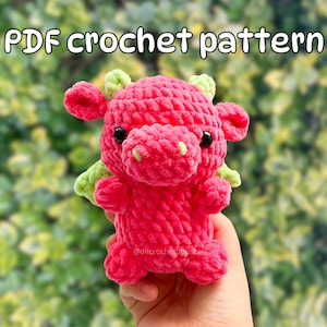 Puede incluir: Patrón de crochet PDF para un amigurumi de dragón rosa y verde. El dragón tiene una cara linda con ojos negros y una pequeña nariz verde.