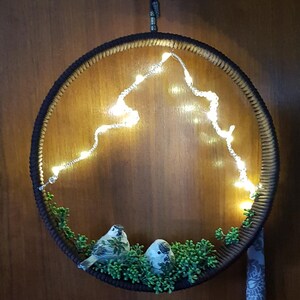 Könnte beinhalten: Runde Wanddekoration mit einem dunkelbraunen, gewebten Rahmen. Im Inneren bilden warmweiße Lichterketten eine bergähnliche Form. Zwei kleine Vogelfiguren sitzen auf grünem Laub. Ein kleiner Stoff mit Muster hängt unten.