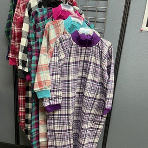 May include: A collection of plaid sleepwear in various colors, including red, black, teal, and purple. The sleepwear is hanging on a metal rack. Each garment has long sleeves and a collared neckline.
