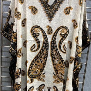 May include: White kaftan with black and gold paisley print. The kaftan has a V-neck and short sleeves. The fabric is sheer and lightweight. The kaftan is made by Metropolitan.
