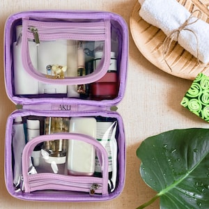 May include: A lavender rectangular toiletry bag with two zippered compartments and clear plastic windows. The interior is organized with various white and clear bottles, tubes, and containers. The brand name AKU is printed on the center divider.