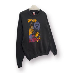 Puede incluir: Sudadera negra de cuello redondo con un gr&aacute;fico de un jugador de baloncesto en amarillo y morado. La palabra "JORDAN" est&aacute; impresa en rojo y amarillo. La sudadera est&aacute; colgada en una percha blanca.