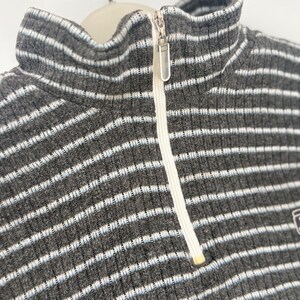 May include: A charcoal gray ribbed sweater featuring a high collar and a quarter-length front zipper. The garment is decorated with thin, horizontal white and light blue stripes throughout, and includes a silver-toned metal zipper pull.