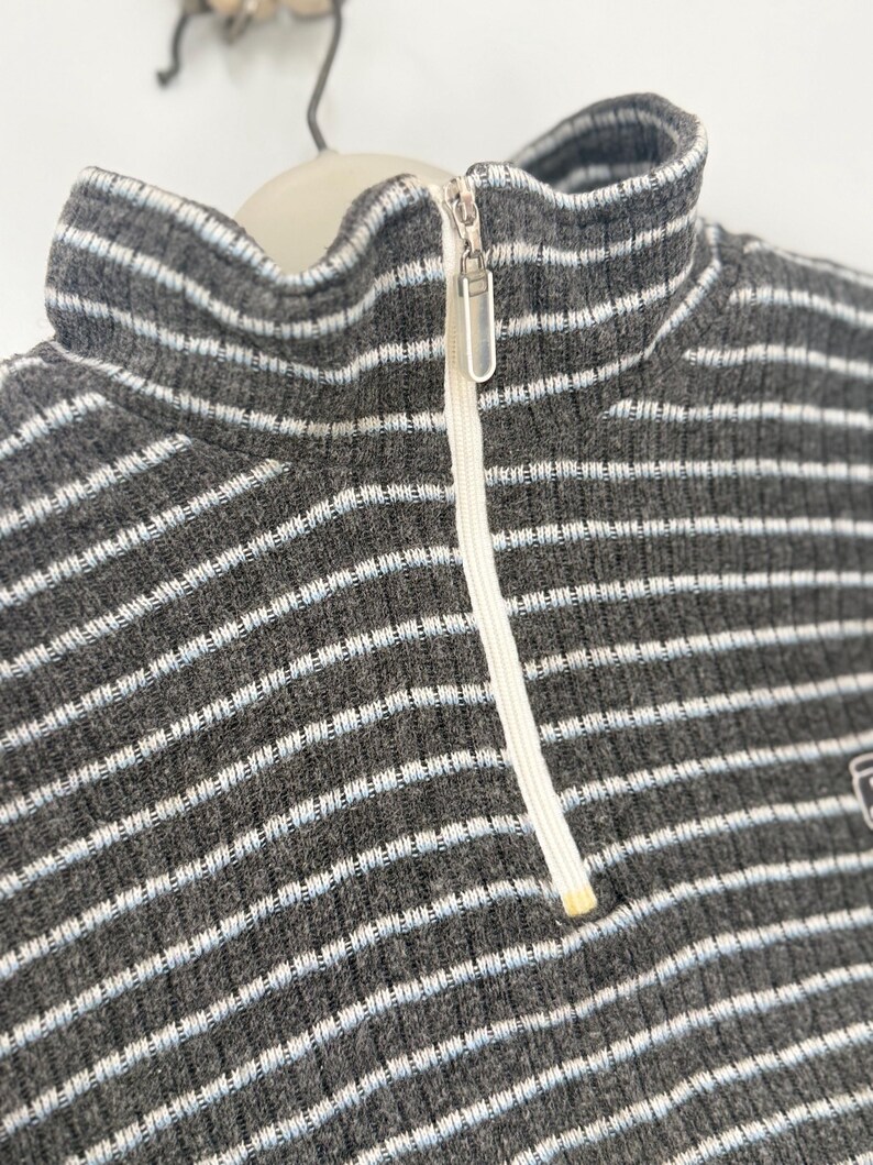 May include: A charcoal gray ribbed sweater featuring a high collar and a quarter-length front zipper. The garment is decorated with thin, horizontal white and light blue stripes throughout, and includes a silver-toned metal zipper pull.
