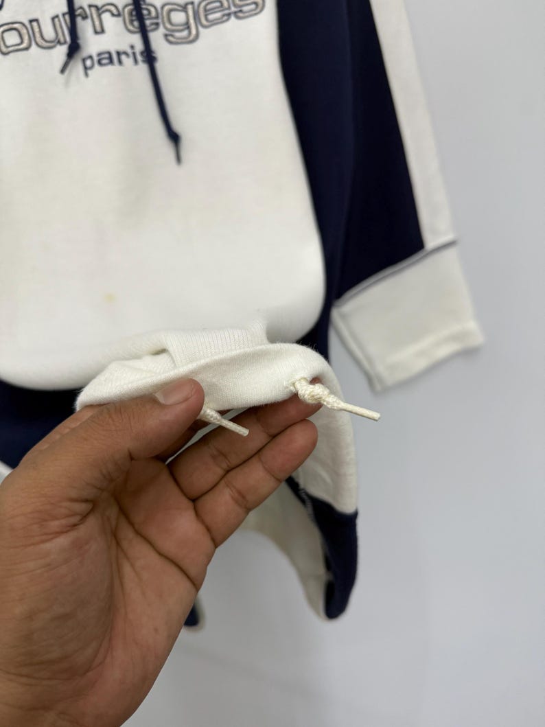 Puede incluir: Sudadera con capucha blanca y azul marino con el texto "courr&egrave;ges paris" en gris. La sudadera tiene un cord&oacute;n y un panel azul marino en contraste en el lateral. Los pu&ntilde;os son blancos.