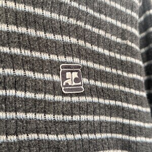 May include: A dark gray ribbed knit sweater featuring horizontal light blue stripes. The garment displays a small, rectangular embroidered patch on the chest, which contains a stylized white logo resembling the letters AC on a dark background.