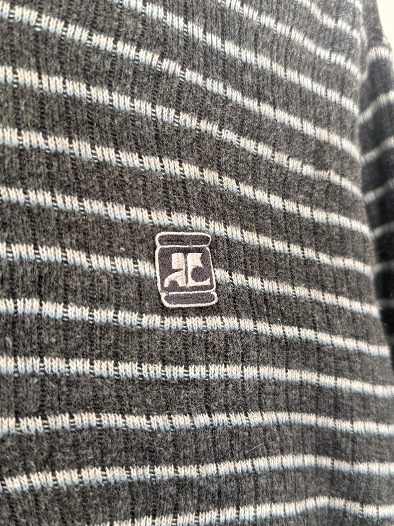 May include: A dark gray ribbed knit sweater featuring horizontal light blue stripes. The garment displays a small, rectangular embroidered patch on the chest, which contains a stylized white logo resembling the letters AC on a dark background.