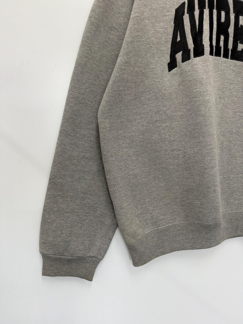 Pode incluir: Camisola cinzenta com a palavra "AVIRE" em letras grandes e pretas no peito. A camisola tem gola redonda e mangas compridas com punhos canelados. O tecido parece ser um material macio e confort&aacute;vel.