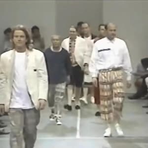 Könnte beinhalten: Eine Modenschau mit mehreren Personen, die über einen Laufsteg gehen. Die Models tragen verschiedene Outfits, darunter Jacken, Hemden und Hosen mit Mustern. Die Farbpalette umfasst Weiß, Blau und Beige.