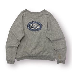 May include: A heather gray sweatshirt with a crew neck. The back features an embroidered oval design with the word "ROXY" in blue and white, surrounded by a floral pattern. The sweatshirt has long sleeves.