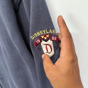 May include: A blue sweatshirt sleeve with an embroidered Disneyland patch. The patch features the word "Disneyland" in gold lettering, the numbers "19" and "55", and a red "D" on a white background.