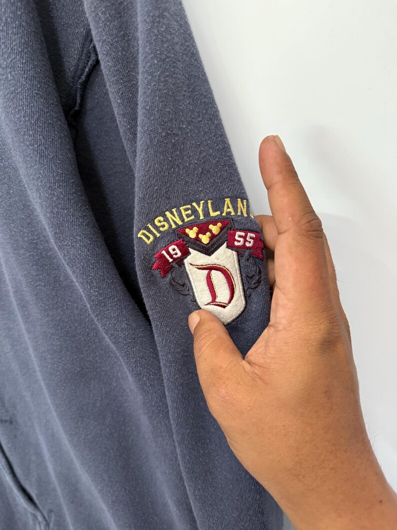 May include: A blue sweatshirt sleeve with an embroidered Disneyland patch. The patch features the word "Disneyland" in gold lettering, the numbers "19" and "55", and a red "D" on a white background.