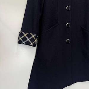 May include: A navy blue coat with a textured fabric. The coat features a classic design with four round, silver-colored buttons and a contrasting plaid cuff detail in yellow, white, and black. The coat has a tailored fit and a subtle A-line shape.