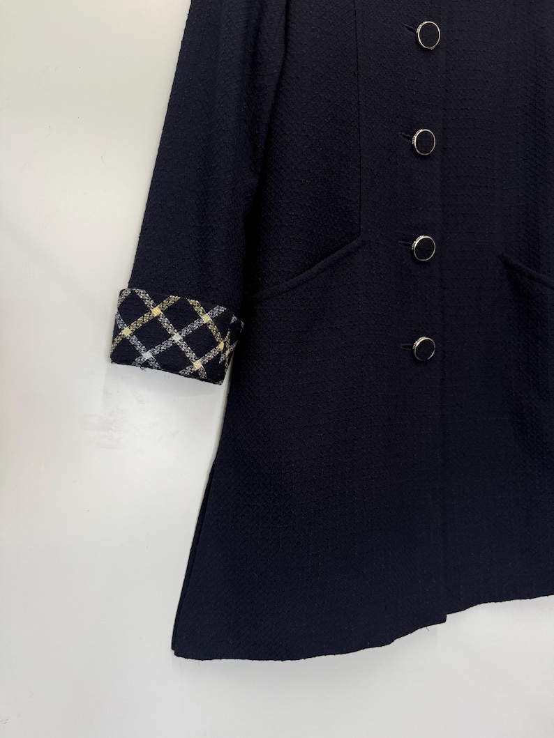 May include: A navy blue coat with a textured fabric. The coat features a classic design with four round, silver-colored buttons and a contrasting plaid cuff detail in yellow, white, and black. The coat has a tailored fit and a subtle A-line shape.