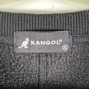 Op de afbeelding: Zwarte fleece trui met een zwart Kangol label met een kangoeroe logo en de tekst "Kangol" in wit. Het label heeft ook een wit "XL" maatlabel.