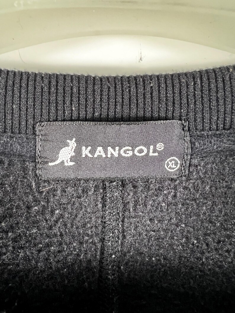 Op de afbeelding: Zwarte fleece trui met een zwart Kangol label met een kangoeroe logo en de tekst "Kangol" in wit. Het label heeft ook een wit "XL" maatlabel.