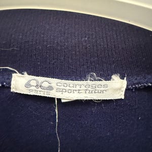 Puede incluir: Primer plano de una prenda azul marino con una etiqueta blanca. La etiqueta presenta el texto "AC courreges sport futur paris" en una fuente estilizada. La tela parece ser un material de punto.