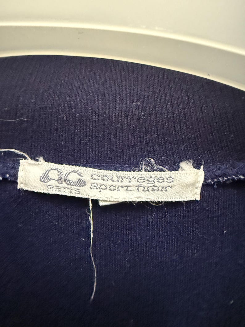 Puede incluir: Primer plano de una prenda azul marino con una etiqueta blanca. La etiqueta presenta el texto "AC courreges sport futur paris" en una fuente estilizada. La tela parece ser un material de punto.