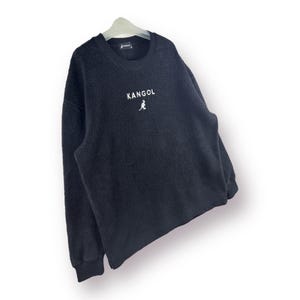 Op de afbeelding: Zwart fleece sweatshirt met een wit kangoeroe logo en het woord "Kangol" gedrukt op de voorkant.